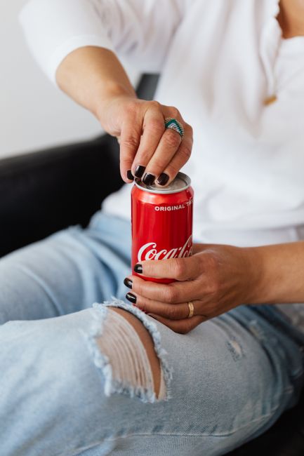 Crop unrecognizable lady in trendy jeans and white blouse with manicure opening red can of coke while sitting on cozy armchair near white wall