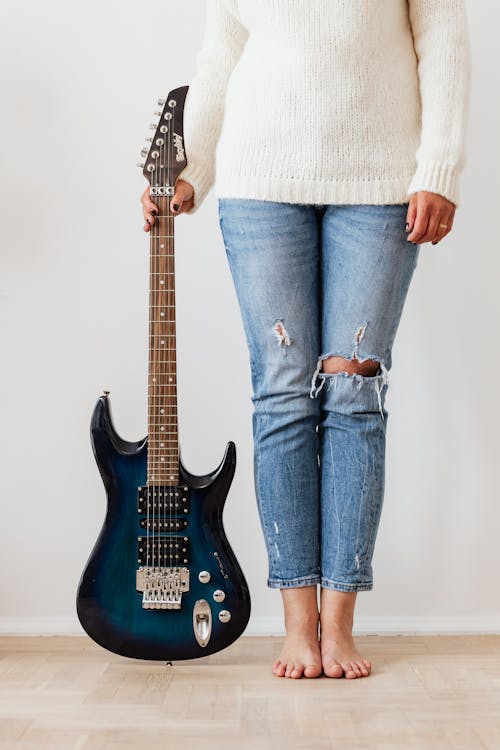 Crop unrecognizable barefoot female in ripped jeans and white knitted sweater standing near electric guitar on parquet against white wall