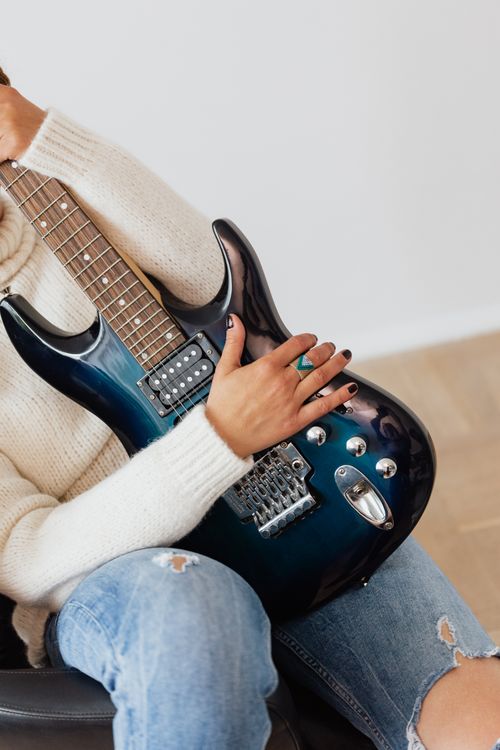 Free Crop unrecognizable female in ripped jeans and white knitted sweater sitting on black chair with electric dark blue guitar on laps Stock Photo