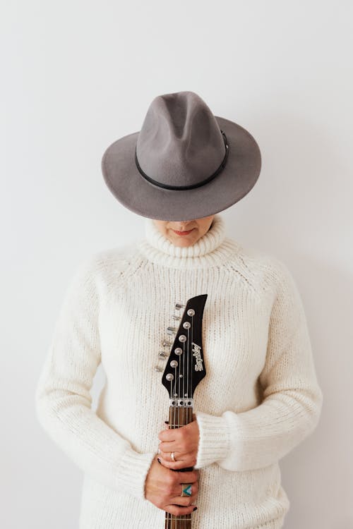 Unrecognizable female musician wearing white knitted sweater and stylish hat standing against white plain background with electric guitar fretboard in hands