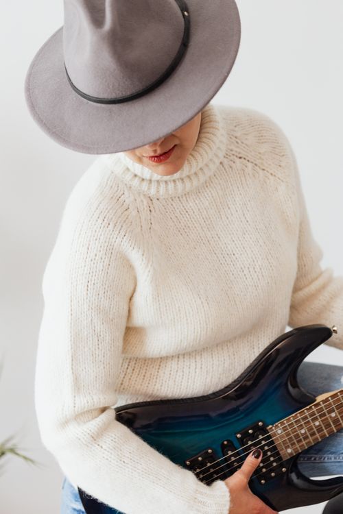 Free Female musician in white knitted sweater and stylish hat performing on modern electric dark blue guitar against white wall Stock Photo