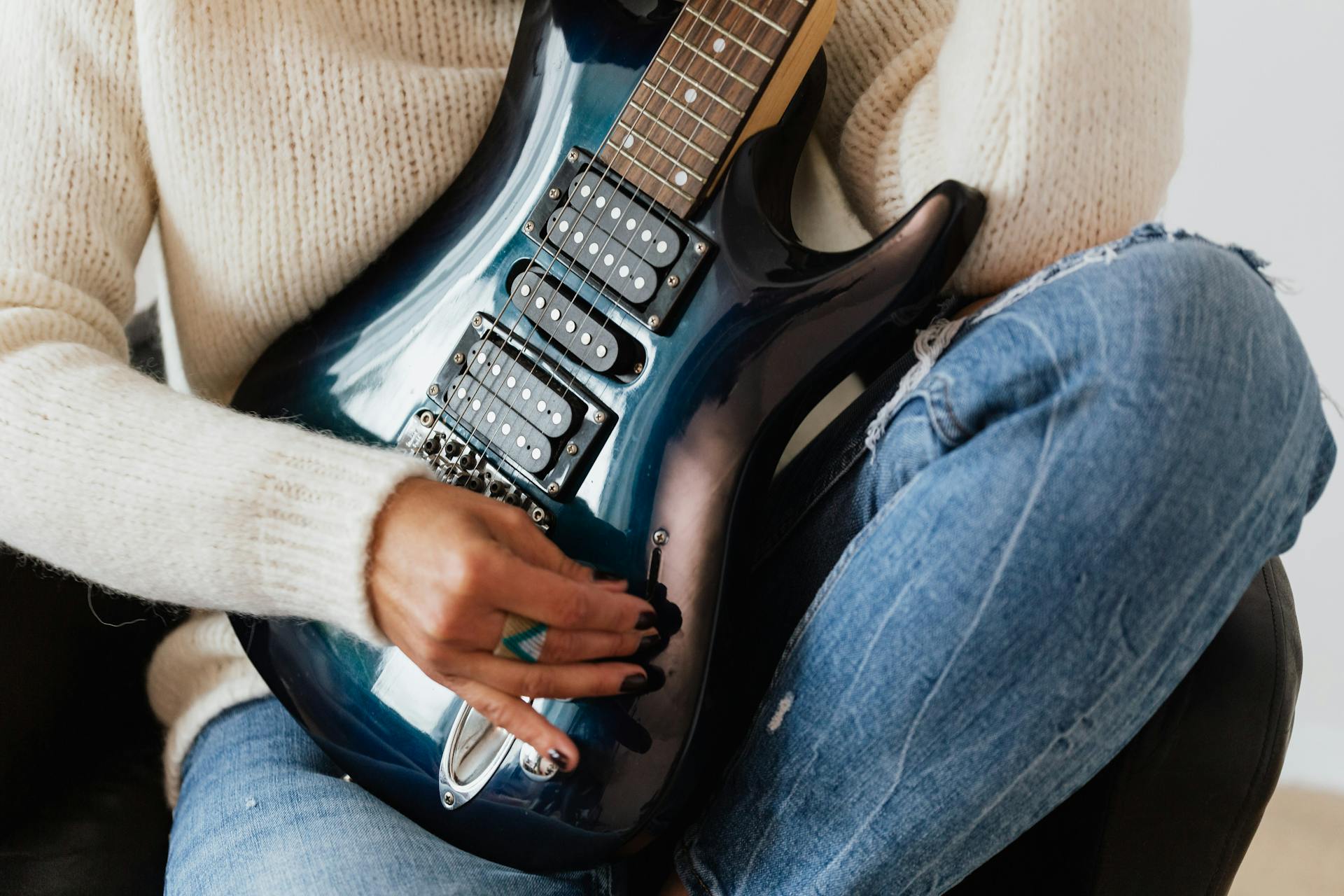 Crop unrecognizable female guitarist wearing jeans and sweater performing electric guitar while sitting on leather chair