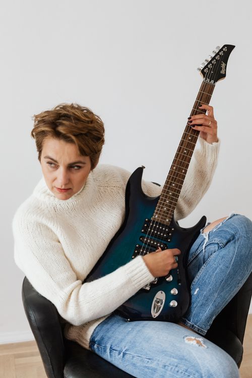Free Positive pensive female musician in jeans and white sweater sitting on black comfy chair and performing electric guitar against white wall Stock Photo
