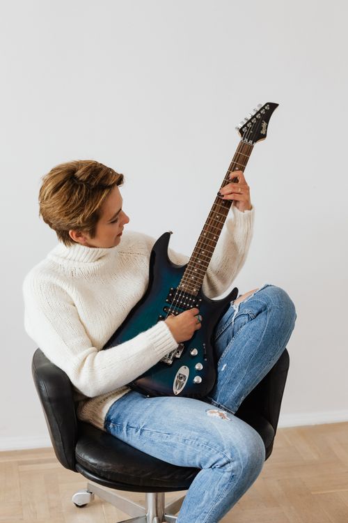 Free Focused female musician wearing jeans and white knitted sweater sitting on black leather chair and performing electric dark blue guitar against white wall Stock Photo