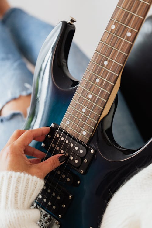 From above of crop anonymous female musician in warm sweater and jeans playing electric guitar while resting in comfortable armchair