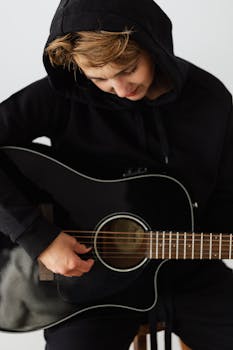 A young musician in a black hoodie strumming an acoustic guitar, focusing intently on the strings.