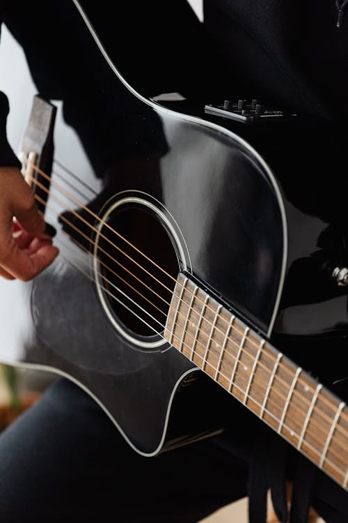 Crop faceless musician in black wear playing classic five string shiny guitar of black color while sitting on stool in apartment