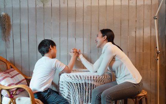 Happy smiling mother and son wearing casual clothes sitting on wooden chairs and competing in arm wrestling in rural house