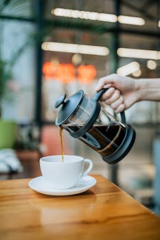 Hand pouring coffee from French press into ceramic cup on wooden table.