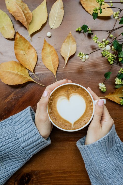 Free A warm latte with heart foam art held by hands, surrounded by autumn leaves on a wooden table. Stock Photo