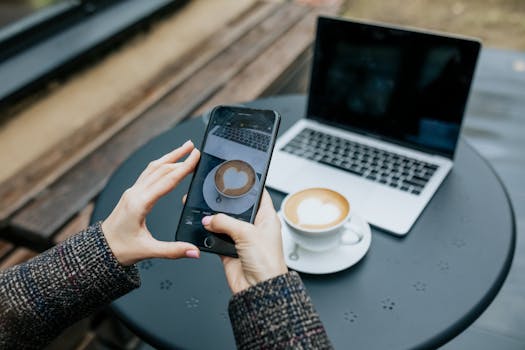 Person snaps a photo of a latte using a smartphone at an outdoor cafe table with a laptop nearby.