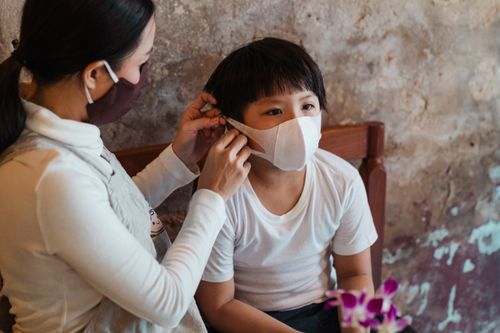 Free Mother wearing white casual wear and protective respirator putting face mask on cute calm son against old shabby weathered wall in daylight Stock Photo