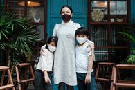 Optimistic Asian woman embracing adorable son and daughter wearing casual clothes and medical protective masks while standing against rural weathered building and plants