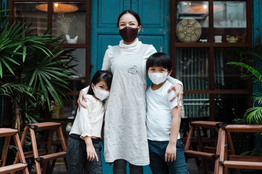 Optimistic Asian woman embracing adorable son and daughter wearing casual clothes and medical protective masks while standing against rural weathered building and plants