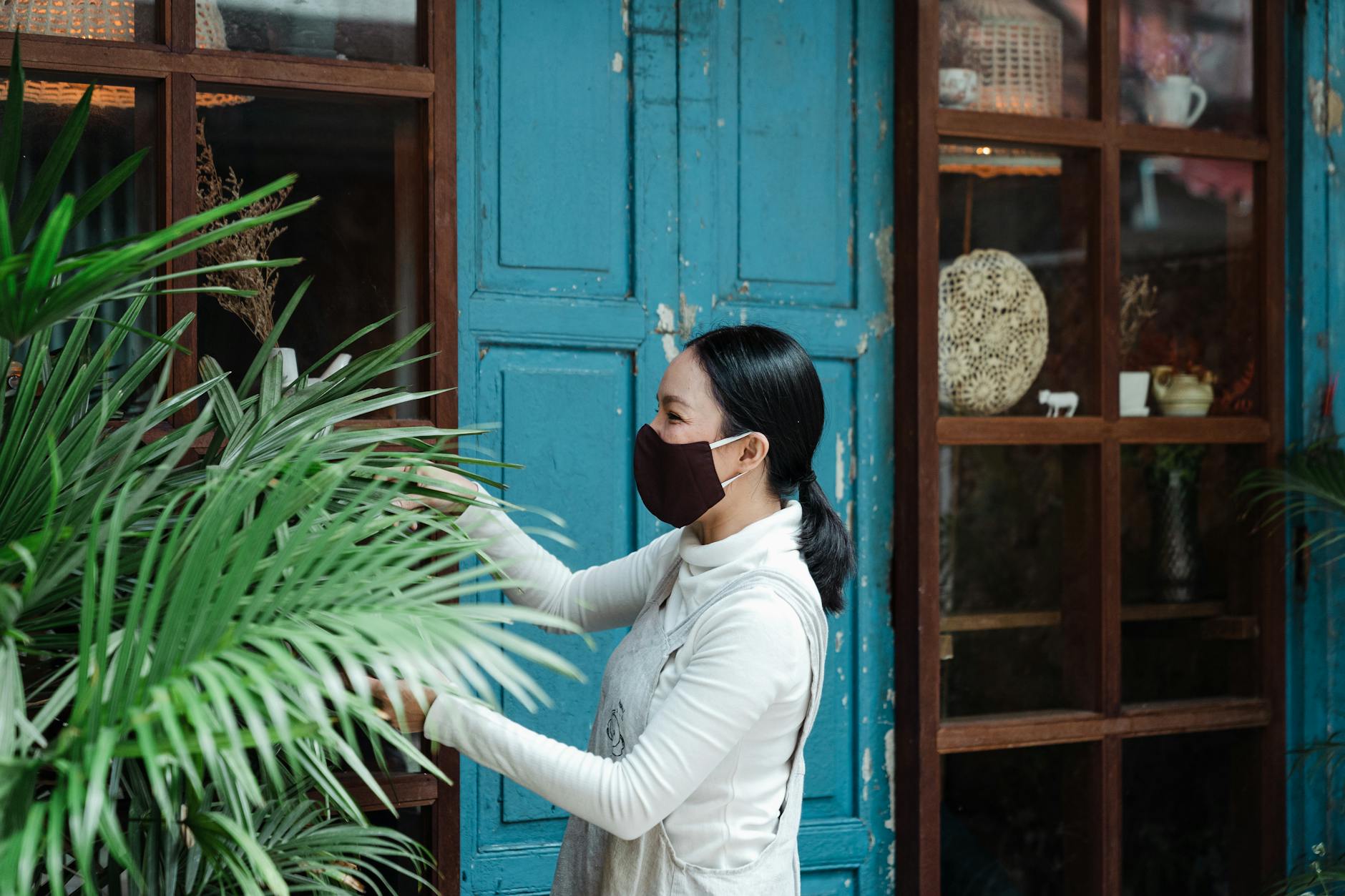 Side view glad Asian lady wearing casual clothes and protective face mask trimming plants in garden near shabby building by using garden scissors