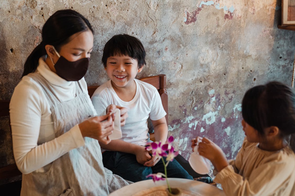Free Optimistic Asian mother in face mask putting respirators on smiling son and daughter while spending time together in rural house against shabby wall Stock Photo