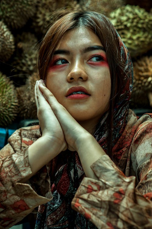 Calm young Asian lady with clasped hands sitting against pile of exotic fruits and looking away