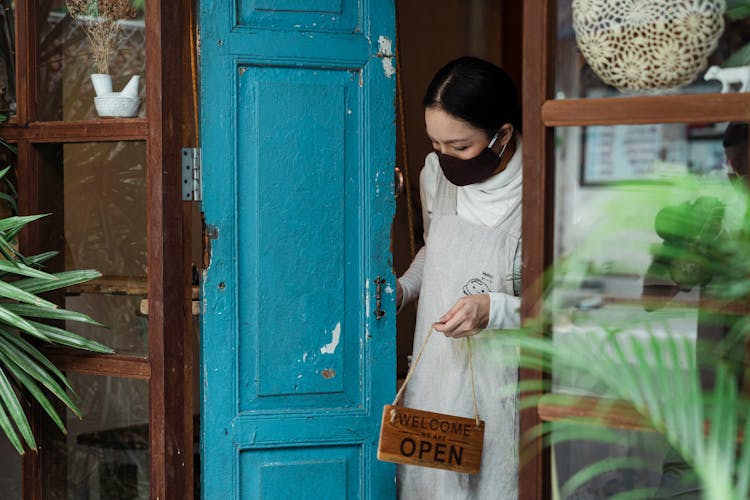 Calm Woman In Respirator With Open Sign Near Shop Door