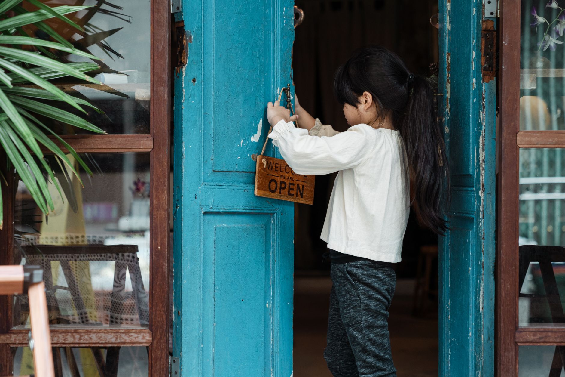 A young girl places an open sign on a rustic wooden door, emphasizing innocence and business.