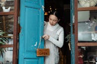 一名妇女将一家咖啡馆蓝色木质门上的“营业中”标志翻转为“欢迎光临”，邀请顾客入内