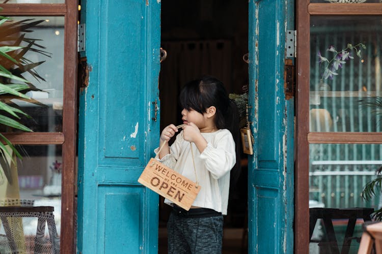 Photo Of Girl Holding Wooden Welcome Signage While Standing On Doorway