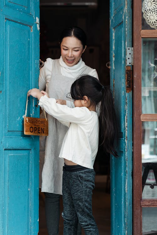 Positive smiling Asian woman with cute daughter in casual wear hanging open sign on blue weathered aged door of rural shop