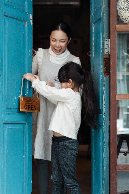 Positive smiling Asian woman with cute daughter in casual wear hanging open sign on blue weathered aged door of rural shop