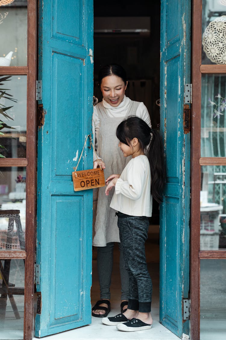 Cheerful Mother And Daughter Hanging Open Sign On Cafe