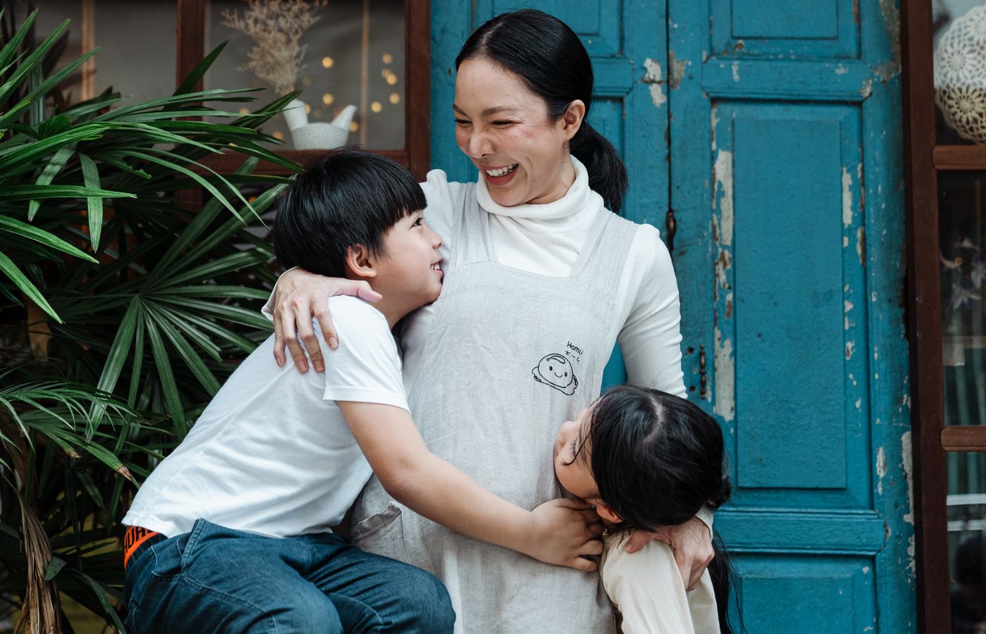 Mother joyfully hugs her son and daughter in front of a rustic blue door, surrounded by greenery.