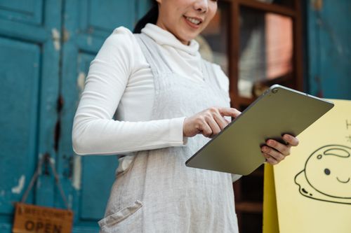 Free Smiling woman with a tablet in front of a shop displaying an open sign. Stock Photo