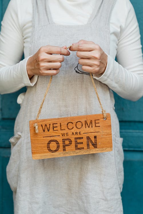 Free Crop faceless female in casual clothes and apron demonstrating wooden sign with text welcome we are open against blue door Stock Photo