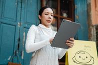 Positive Asian lady in white shirt and gray apron using modern tablet while standing near rural cafe door on daytime
