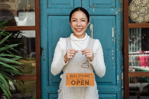 Free Cheerful ethnic female cafeteria owner in apron demonstrating cardboard signboard while standing near blue shabby door and windows after starting own business and looking at camera Stock Photo