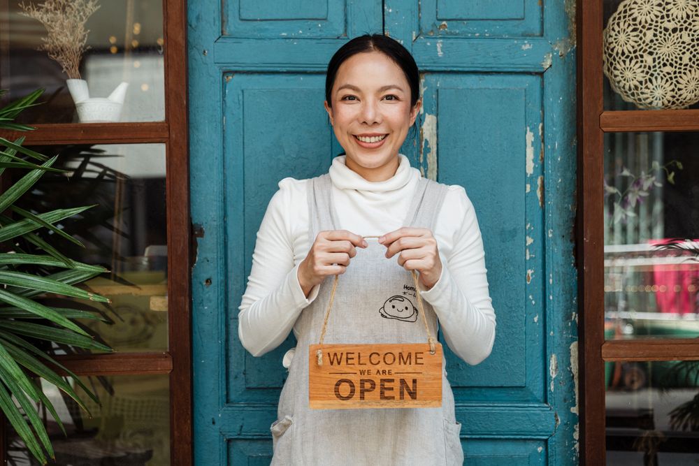 A cafe owner in an apron holding a welcome, we are open sign