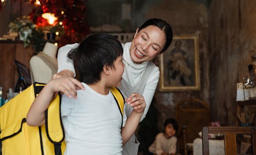 Free Cheerful ethnic female in apron leaned forward while putting on yellow food thermo bag on shoulders of boy in cafeteria with pictures on wall Stock Photo