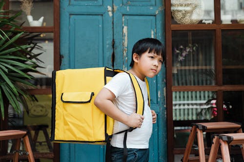 Ethnic boy with heavy food backpack near shabby entrance door