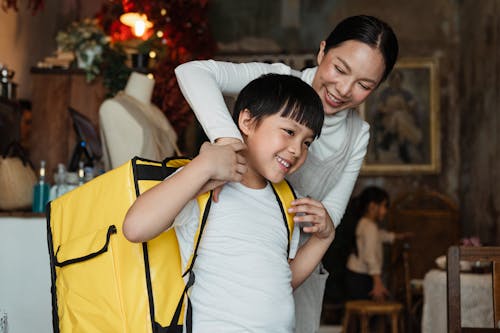 Happy female cafeteria employee putting on big yellow thermo bag on shoulders of positive child in casual wear working as food courier and looking away