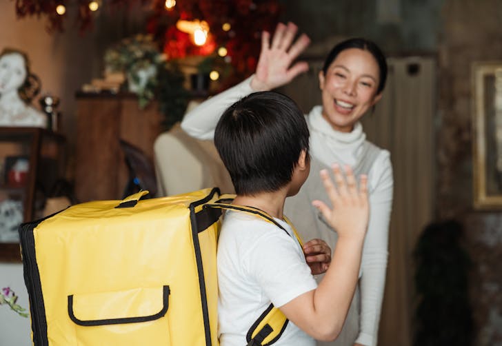 Happy ethnic female cafe worker in apron wishing farewell to boy with food delivery backpack helping in family business