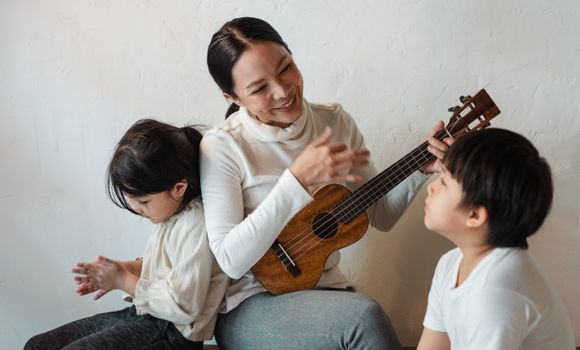 Cheerful ethnic woman in casual wear sitting with crossed legs while playing Hawaiian musical instrument for siblings listening attentively to music