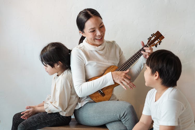 Positive Ethnic Mother Playing Ukulele For Siblings In House