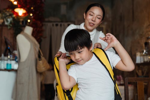 Cheerful ethnic woman helping content schoolboy in casual wear putting on yellow rucksack while standing at home decorated with garland and looking away