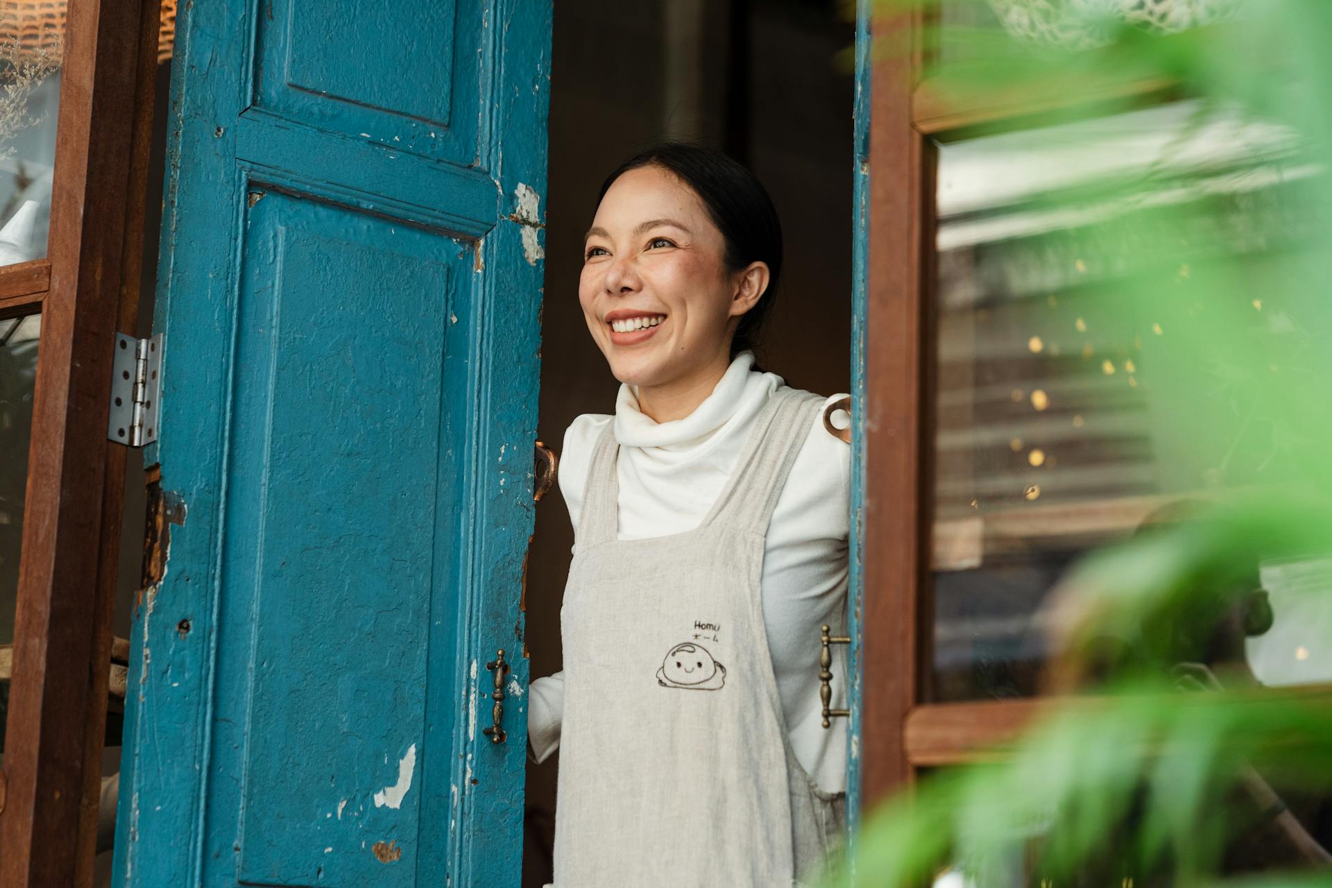 Happy ethnic woman in apron standing at entrance of own cafe