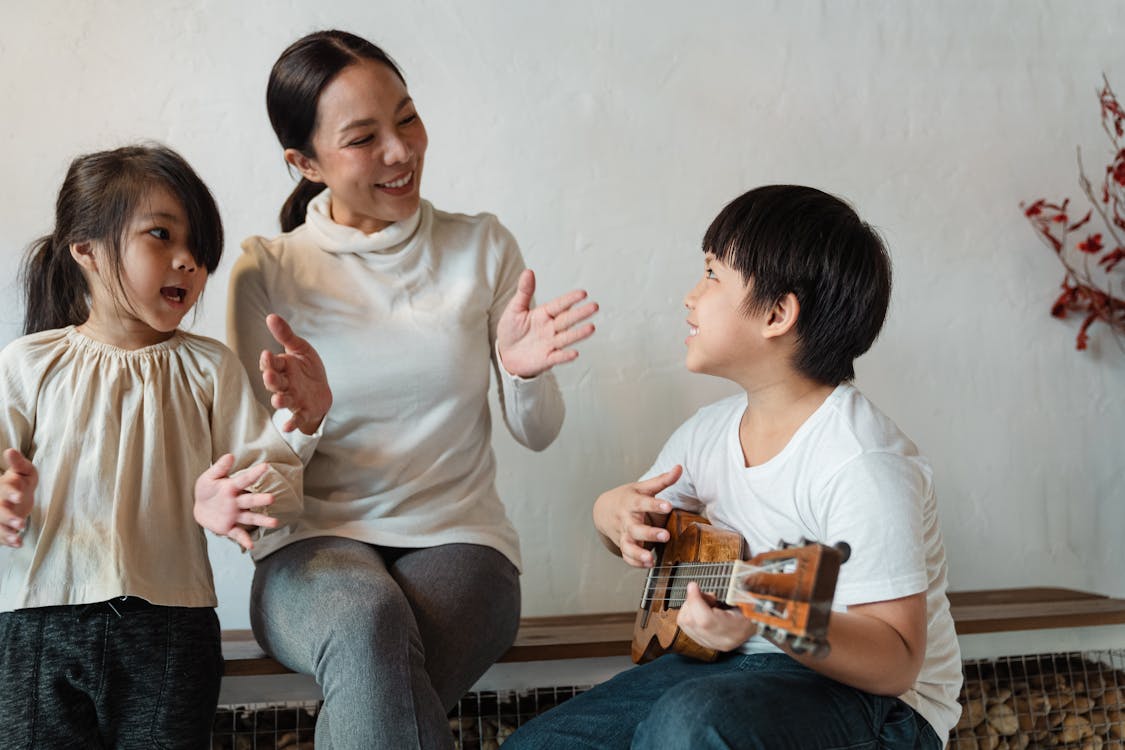 Ethnic boy playing ukulele for cheerful sibling and mother