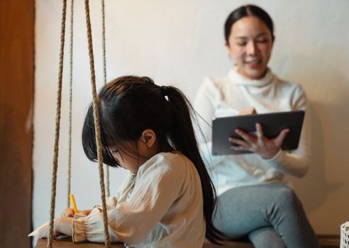 Unrecognizable ethnic girl in casual wear drawing near happy mother sitting with crossed legs while browsing internet on tablet at home