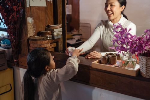 Free Crop cheerful woman in casual wear serving anonymous little girl drink in plastic glass while standing in cafe kitchen near vase of colorful flowers Stock Photo