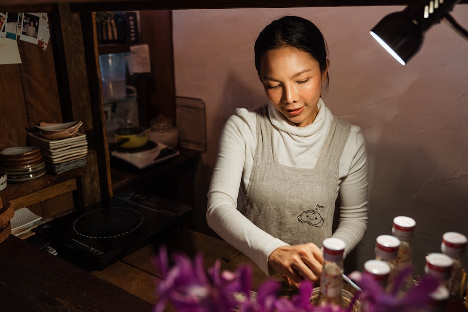 From above of ethnic female worker wearing apron standing behind counter with bottles and looking down while working in cafe with rustic wooden interior in dark light