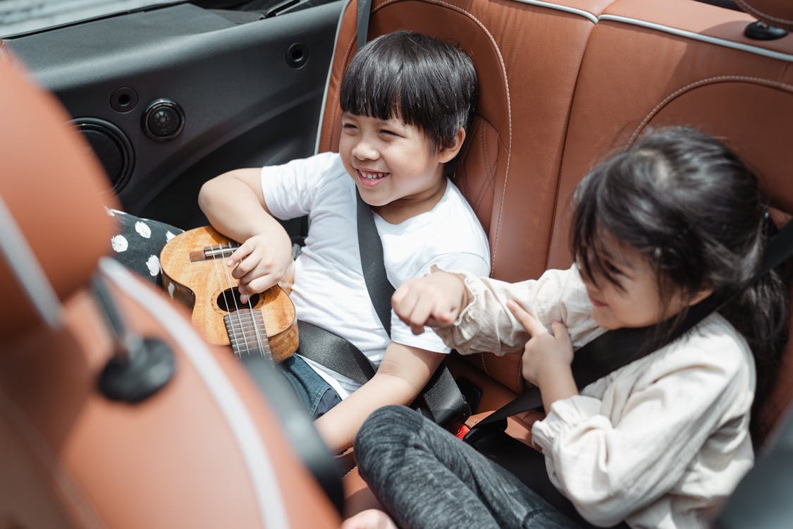 Free From above smiling ethnic boy and girl in casual outfits sitting fastened in passenger seats with ukulele during road trip together Stock Photo