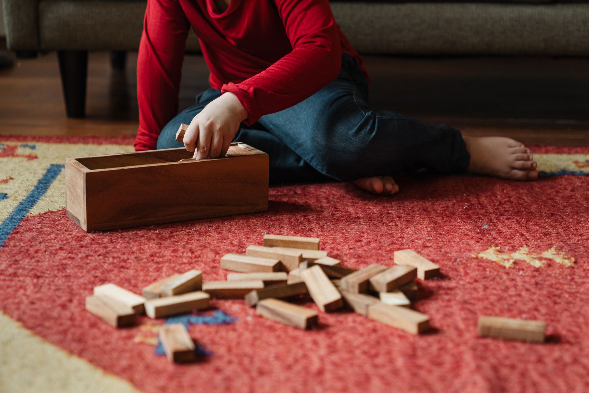 From above crop child in casual clothes sitting on floor carpet near cozy sofa while playing in jenga tower game with wooden blocks improving mental skills
