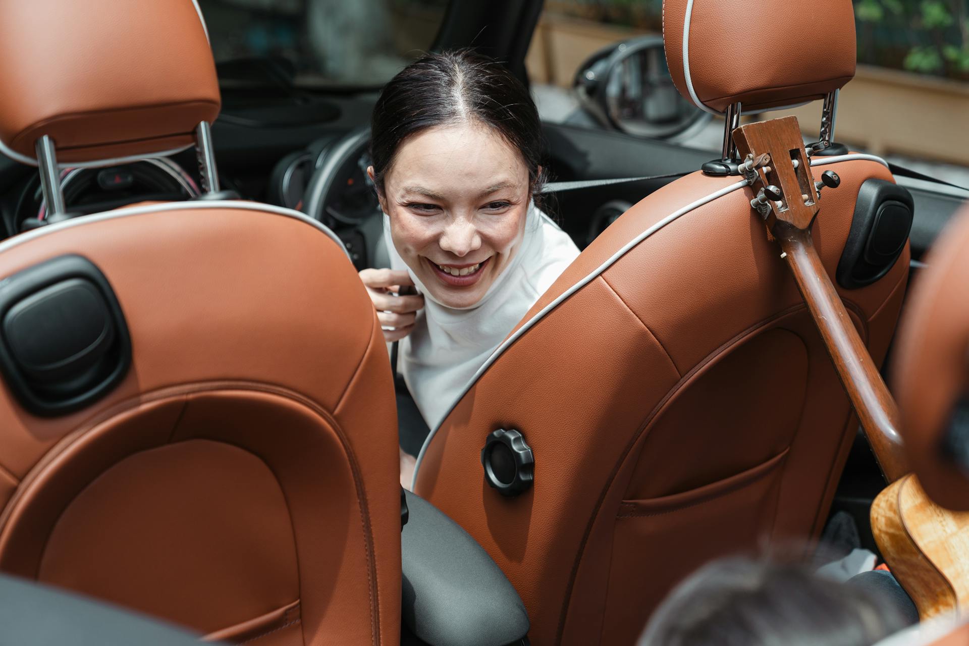 From above of smiling ethnic female driver turning back and looking at unrecognizable kid with ukulele during road trip