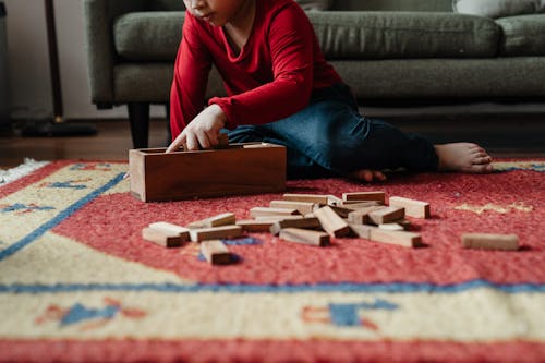 Ground level of cute kid in casual clothes sitting on floor carpet near cozy sofa and playing in jenga tower game with wooden blocks while spending time at home
